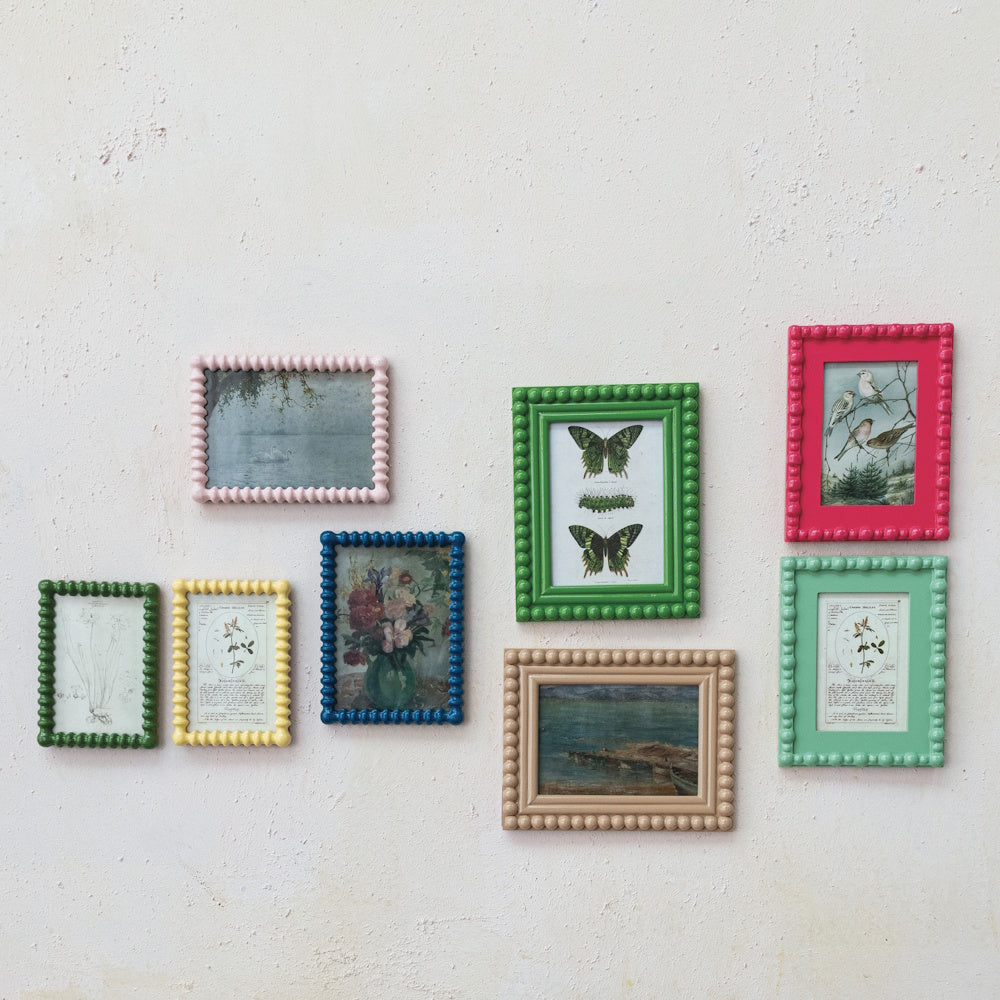 Eight colorful, beaded picture frames holding vintage-style artwork are arranged on a textured white wall.