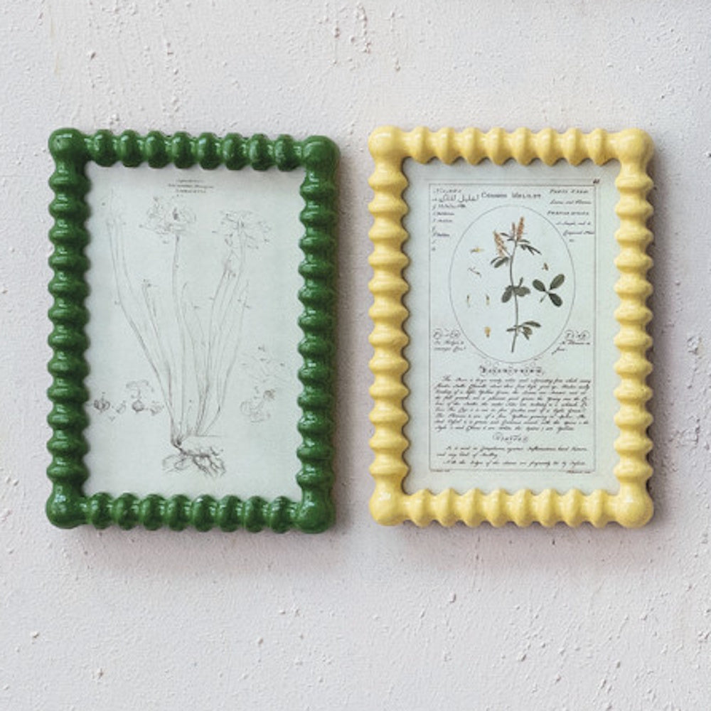Two glossy, wavy-edged picture frames, one green and one yellow, holding vintage botanical prints on a textured wall.