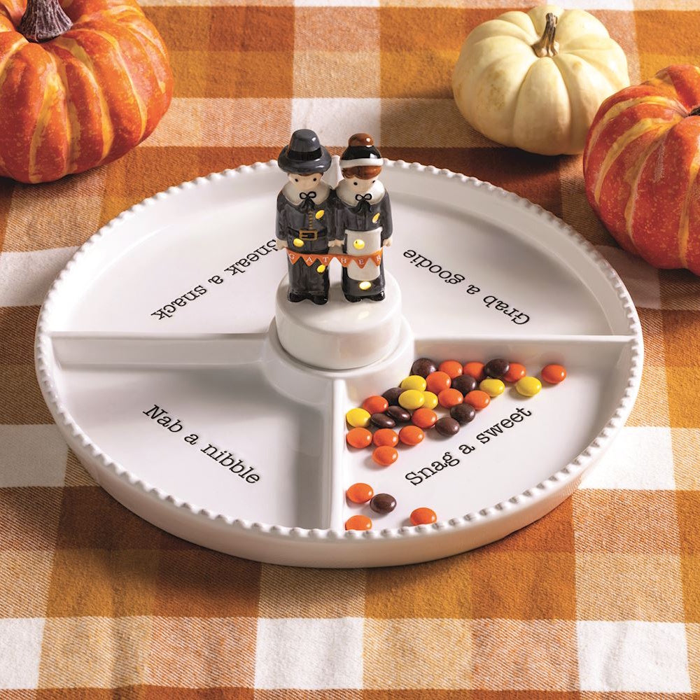 White sectioned snack tray with pilgrim figurines center piece and pumpkins on plaid background.