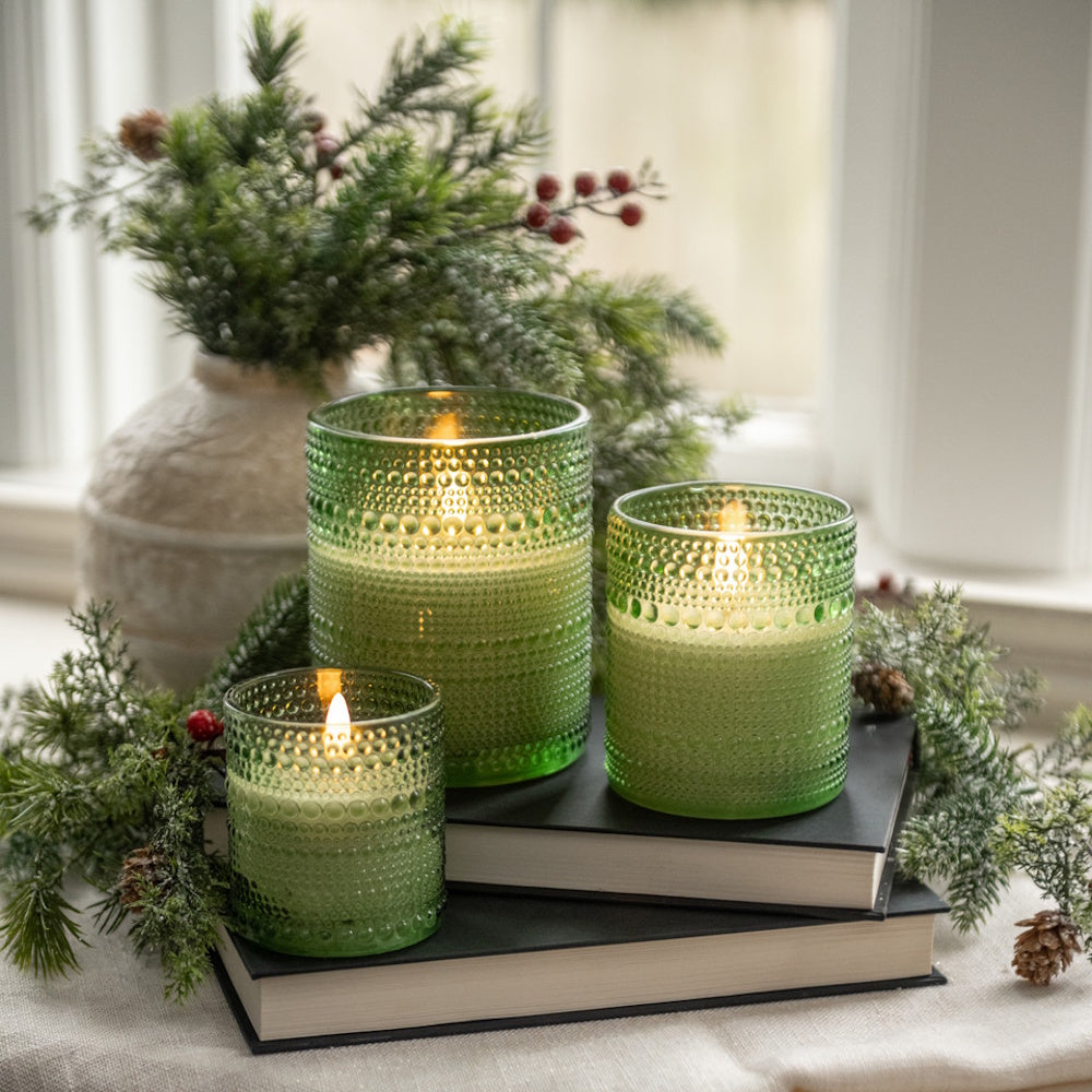 Three lit candles in green dotted glass holders resting on books surrounded by festive pine greenery.