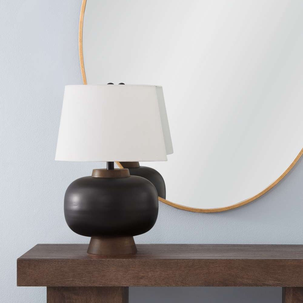 Modern matte black table lamp with white shade on a wooden console table in front of a round gold wall mirror.
