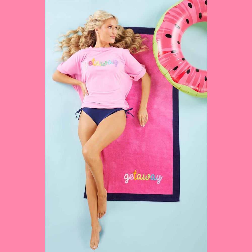 Woman lying on a pink "Getaway" beach towel with a navy border next to a watermelon pool float.