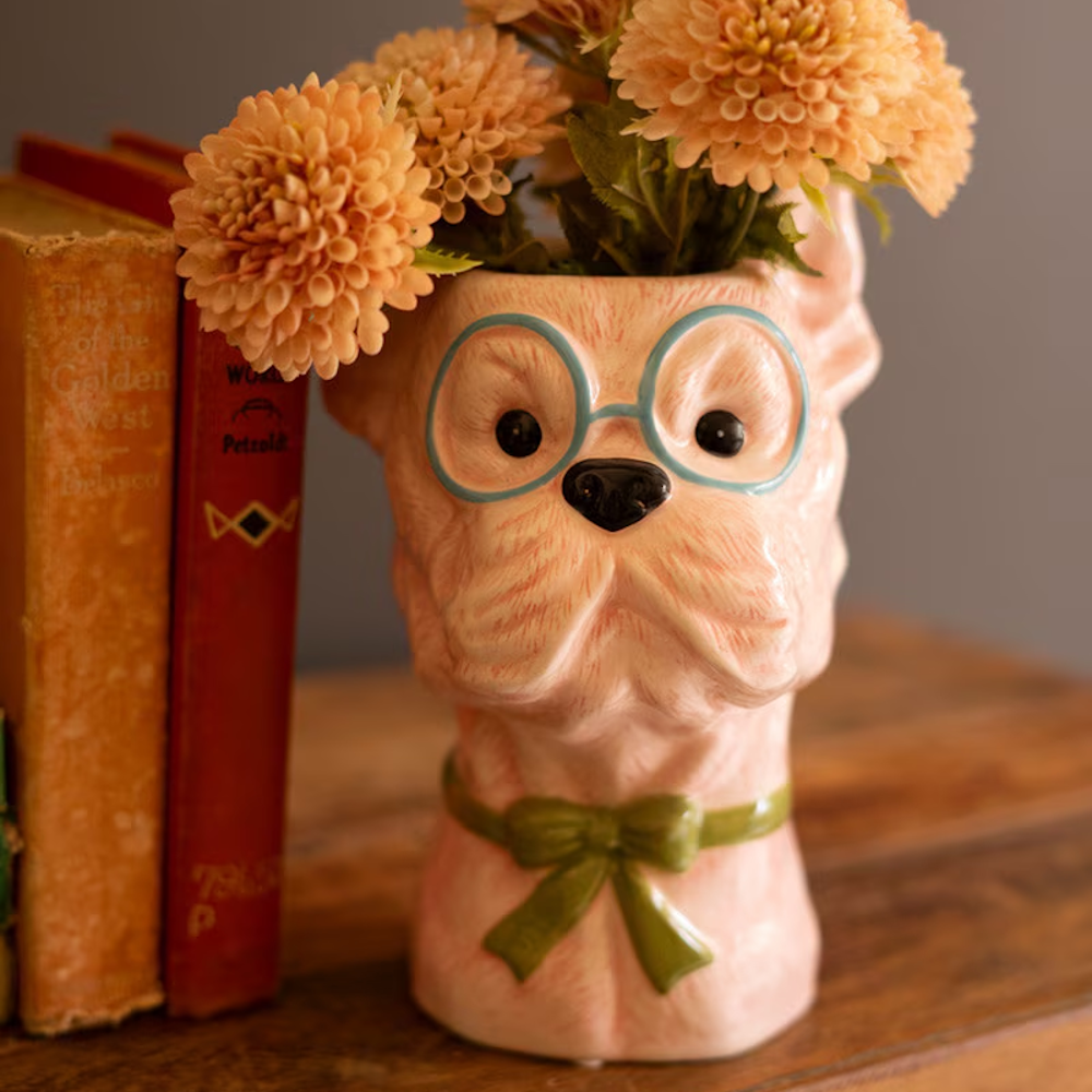 Pink ceramic dog head vase with blue glasses and a green bow, holding orange flowers next to vintage books.
