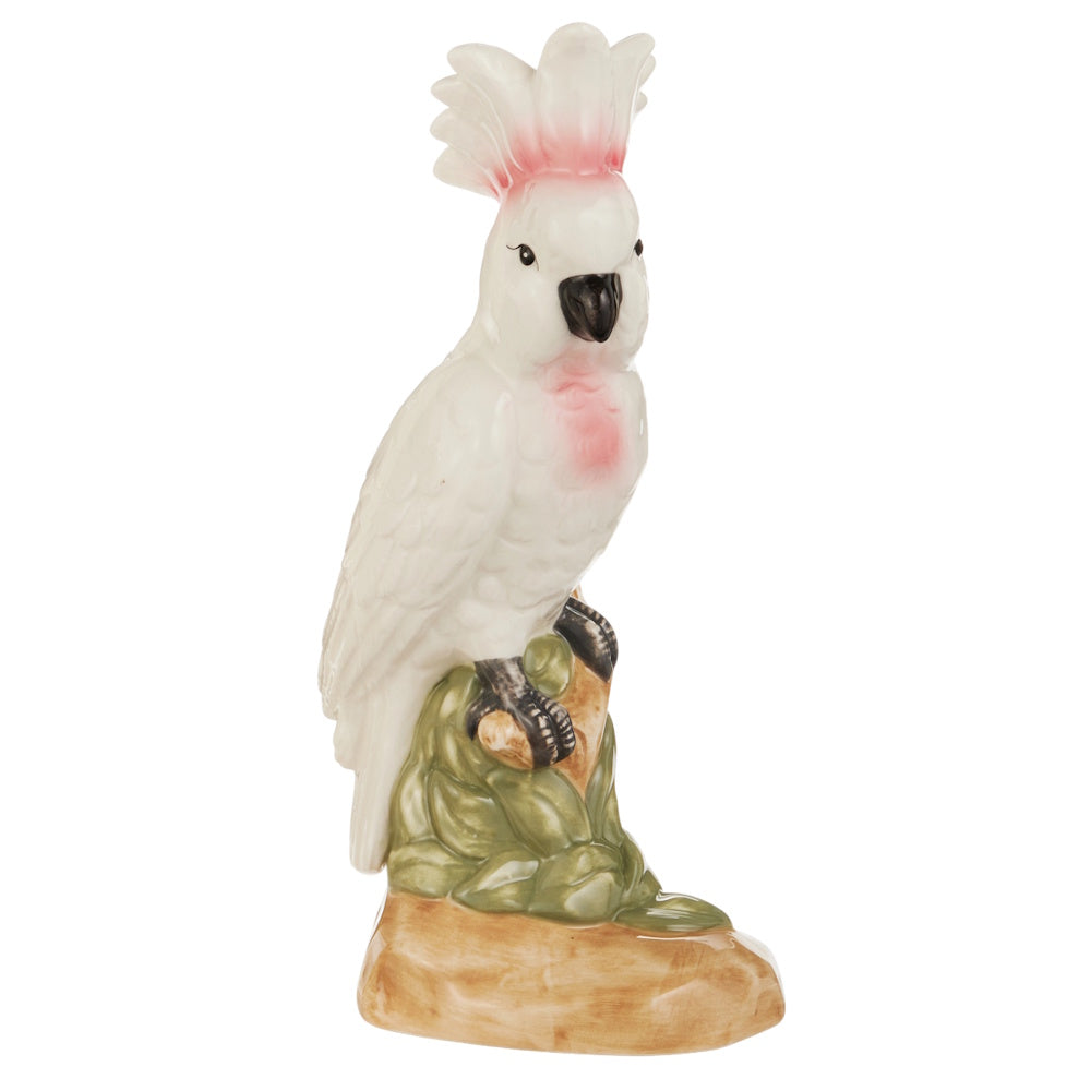 Ceramic figurine of a white cockatoo with pink accents on its crest and chest. The bird perches on a detailed green and brown base.