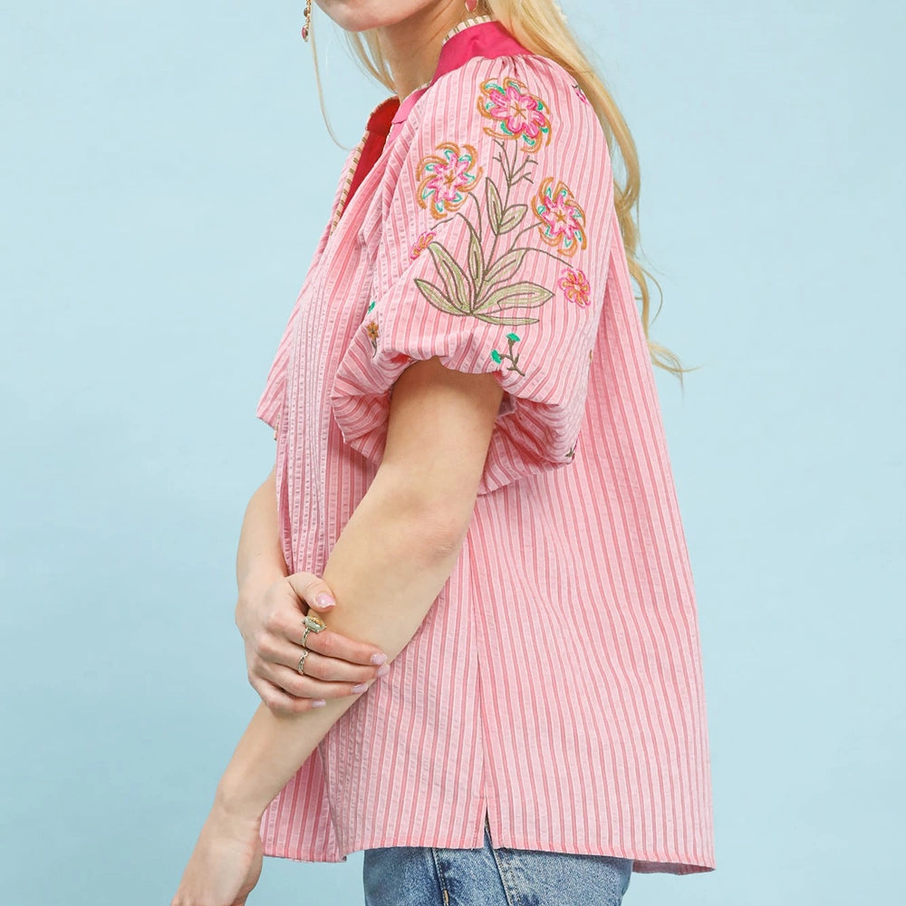 Woman wearing a pink striped puff-sleeve top with floral embroidery and a red collar, paired with blue jeans.