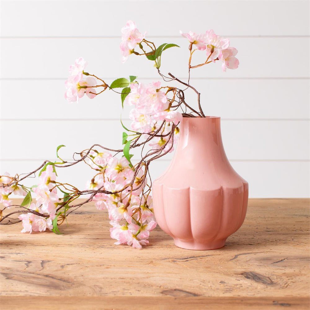 Pink fluted ceramic vase with cherry blossom branches on a rustic wood tabletop. Elegant spring home decor.
