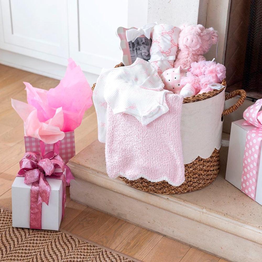 Pink baby girl gift basket with plush toys, blankets, and wrapped presents by a fireplace.