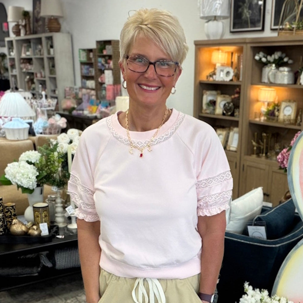 Woman with short blonde hair and glasses wearing a light pink top with lace detail on the collar and sleeves.