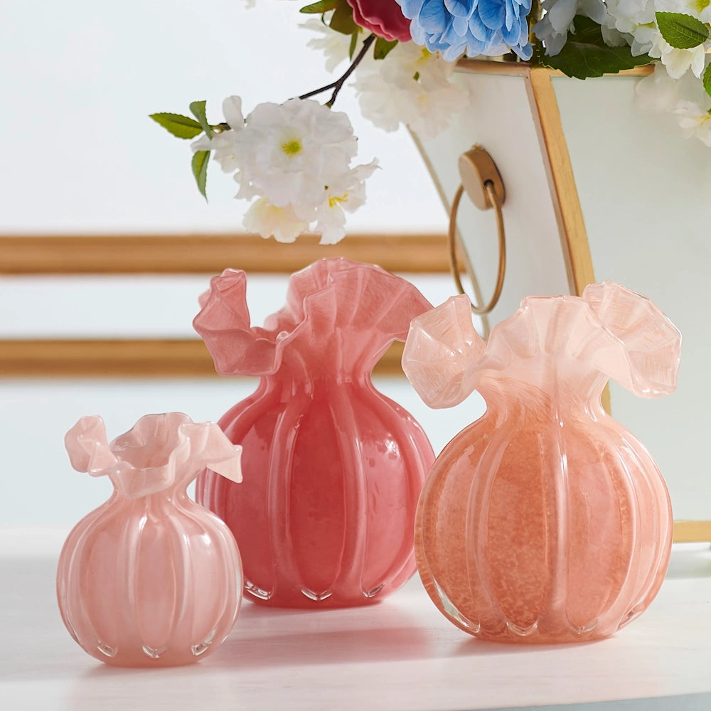 Three pink, ribbed glass vases with ruffled tops in varying sizes and shades arranged on a white surface with flowers.