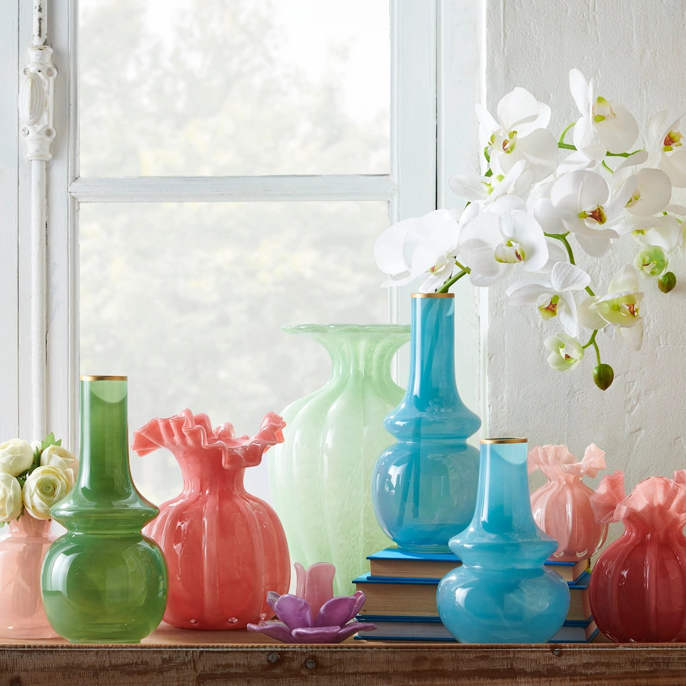 Colorful glass vases in various shapes and shades of pink, blue, and green arranged on a windowsill with white orchids.