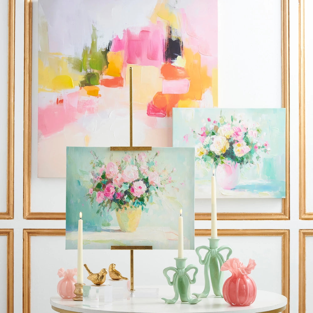 Pastel floral paintings and abstract art displayed on a wall behind a table with candles, gold birds, and pink vases.
