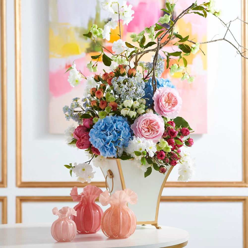 A large floral arrangement in a white and gold vase sits behind three small pink ruffled glass vases on a white table.