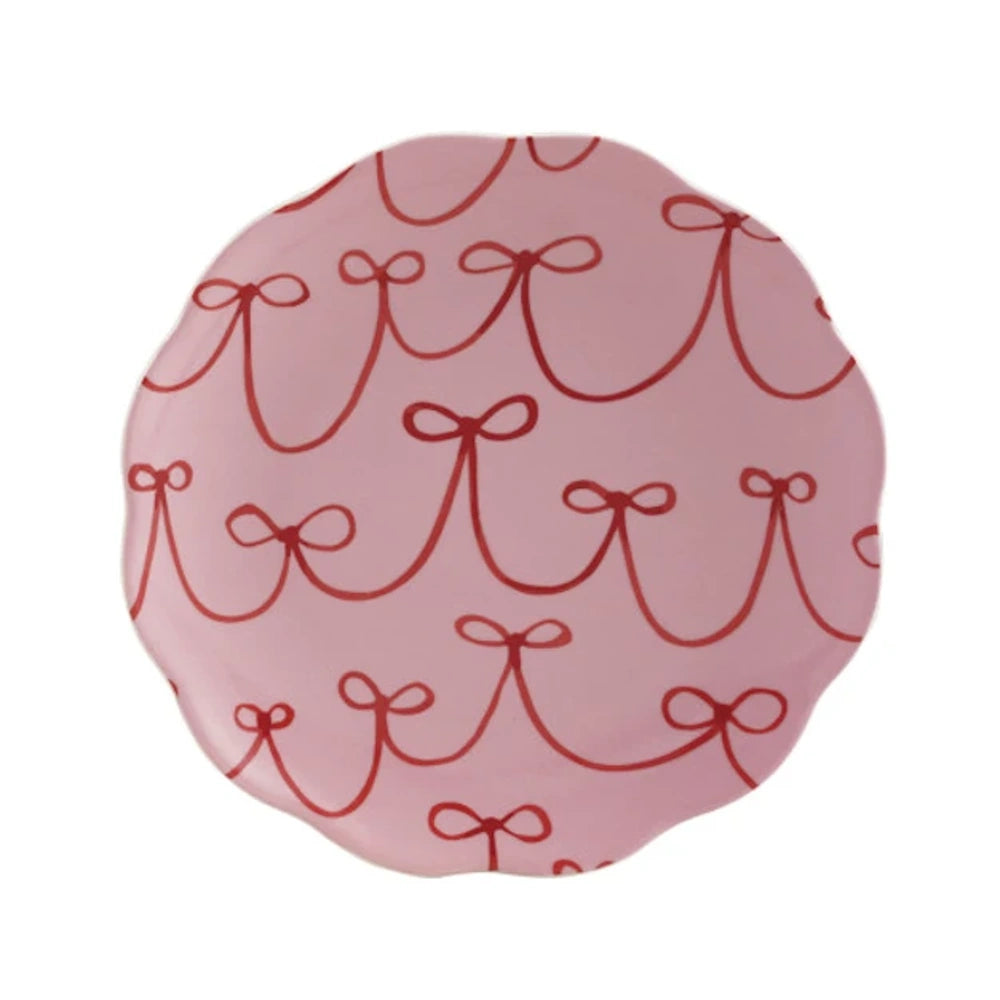 Light pink round scalloped ceramic plate featuring a patterned design of red bows and looping ribbons.