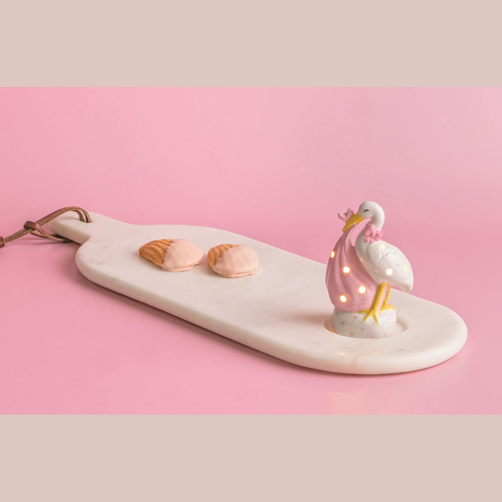 Light-up stork sitter with a pink baby bundle on a marble serving board next to two pink frosted cookies.