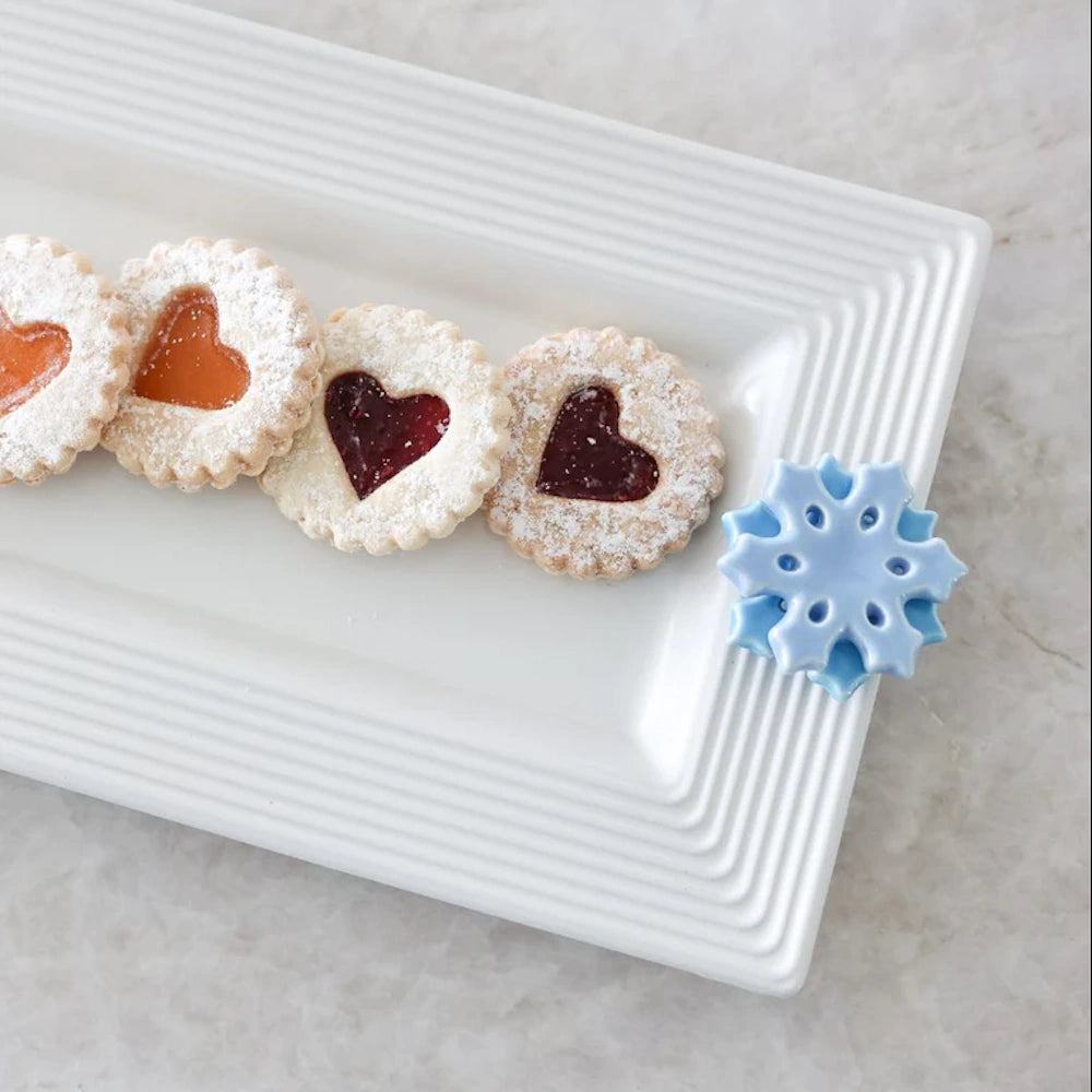 A long white ceramic serving tray holding heart-shaped Linzer cookies, adorned with a blue snowflake charm.