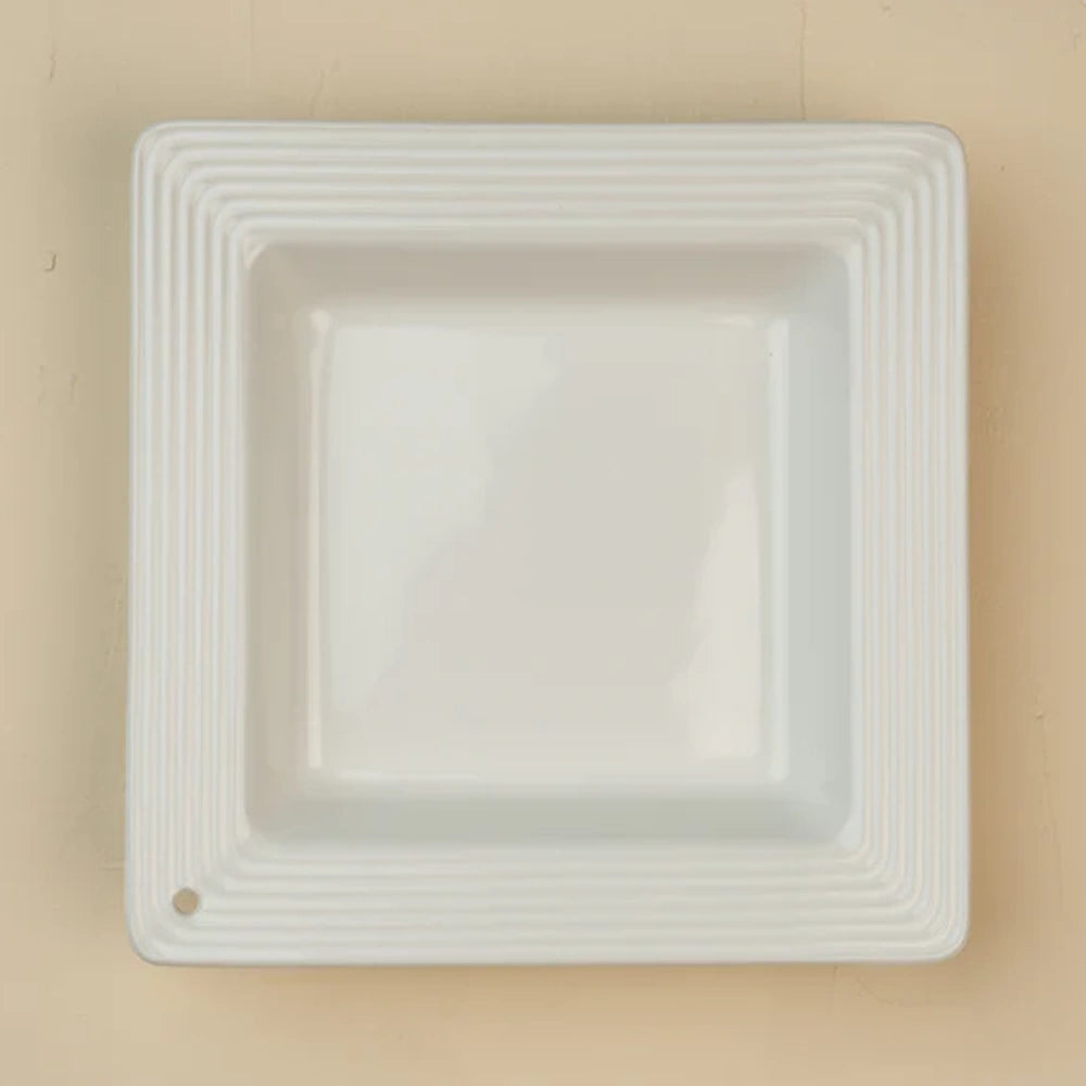 A square white ceramic dish with ridged edges and a single small hole in the bottom left corner.