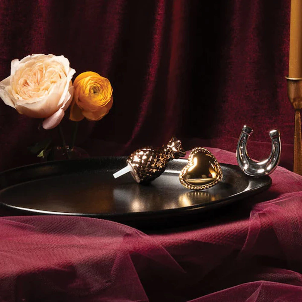 Black oval platter with gold and silver charms set against dark red velvet fabric and roses.