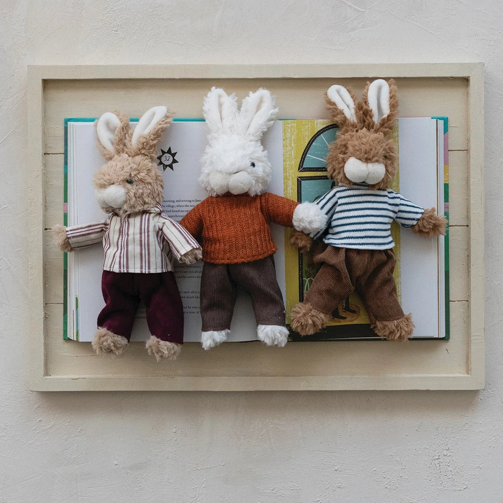 Three plush bunny toys wearing sweaters and pants, posed on an open book inside a wooden frame for nursery room decor.