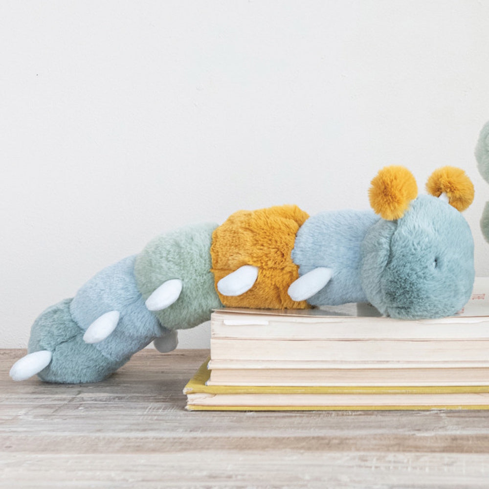 Plush blue, green, and yellow caterpillar toy resting on a stack of books against a white wall.