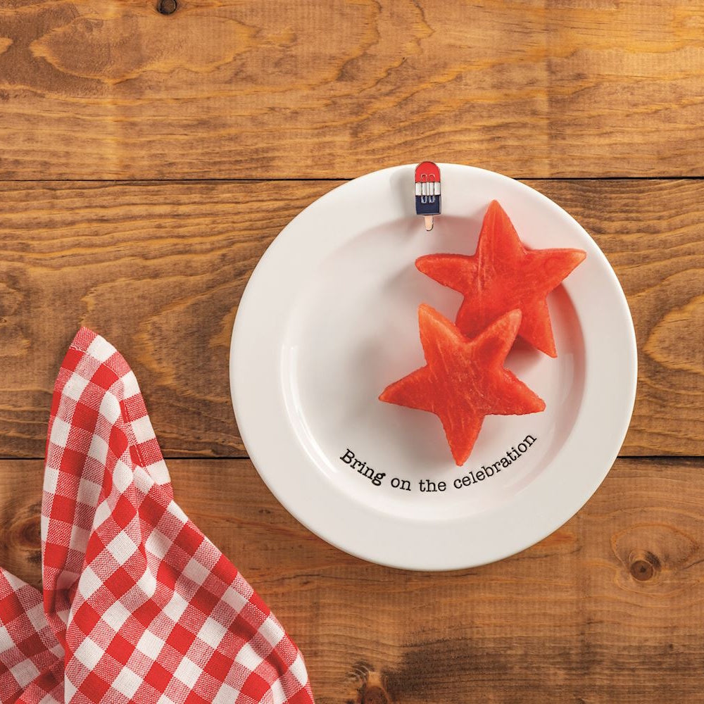 Star-shaped watermelon on a white plate with "Bring on the celebration" text, popsicle charm, and red gingham napkin.