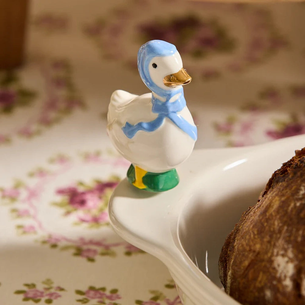 Small ceramic goose figurine wearing a blue bonnet perched on the rim of a white ceramic baking dish.