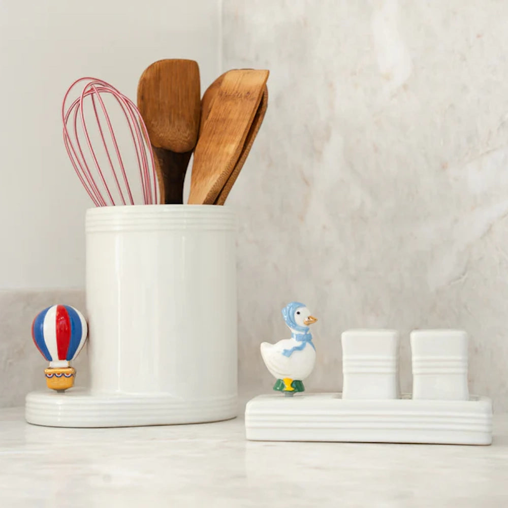 White ceramic utensil crock with spatulas, whisk, and salt and pepper shaker set on a marble countertop.