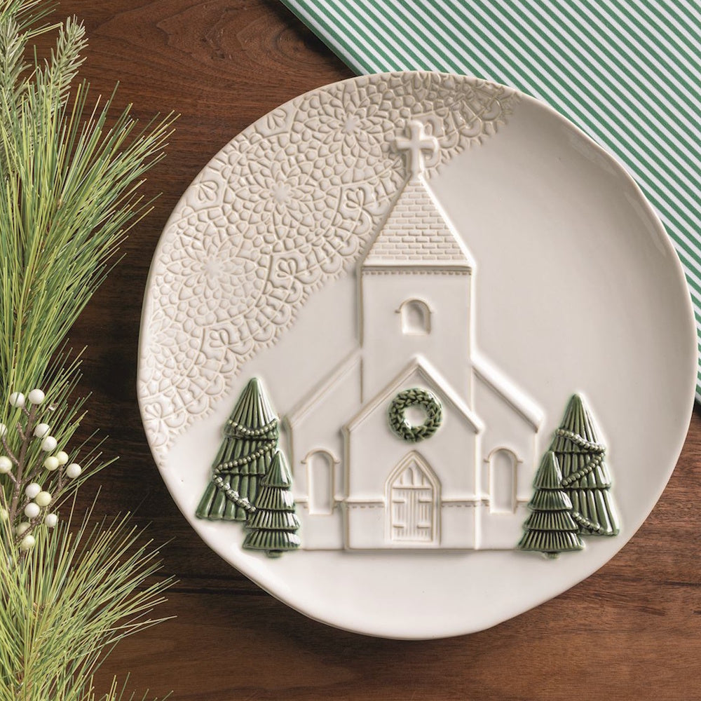 White ceramic platter with embossed church on wooden table beside pine greenery and green striped napkin.