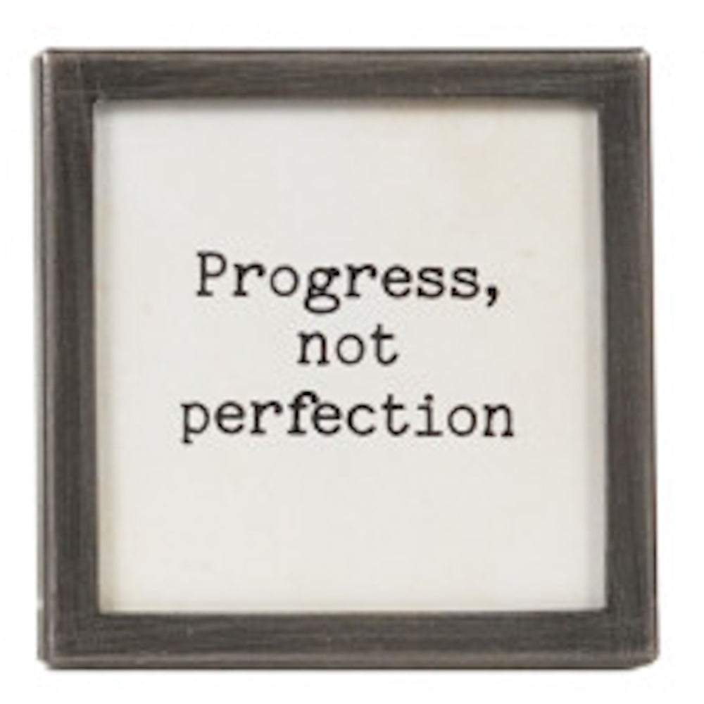A square silver-framed sign with the text "Progress, not perfection" in black typewriter font.