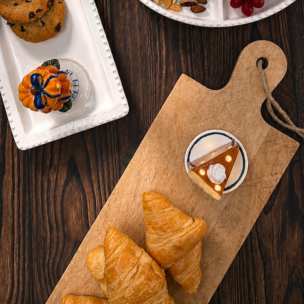 Light-up pumpkin pie slice figurine on a wooden board surrounded by baked pastries and cookies.