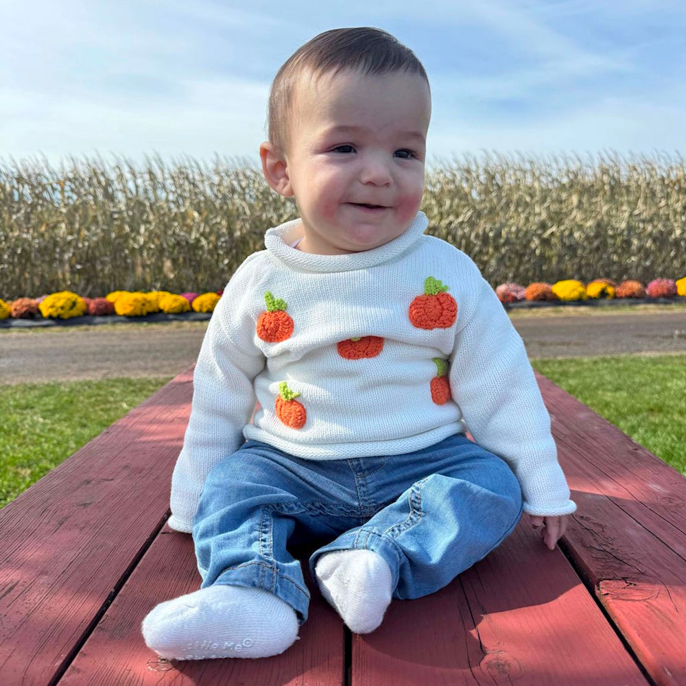 Smiling baby sitting on a red wooden table, wearing a white sweater with knit pumpkins and blue jeans.
