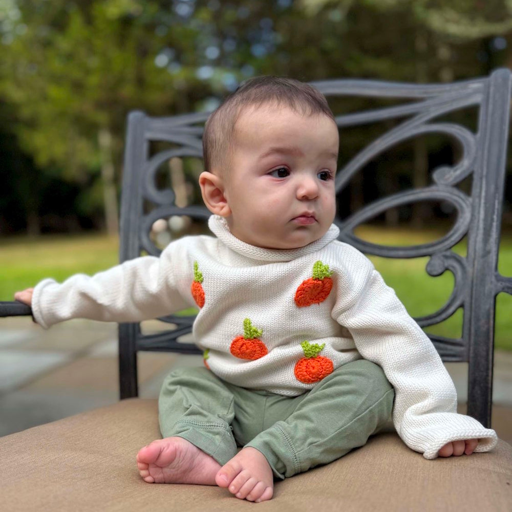 Baby sitting on a chair wearing a white rollneck knit sweater with orange pumpkins and green pants.