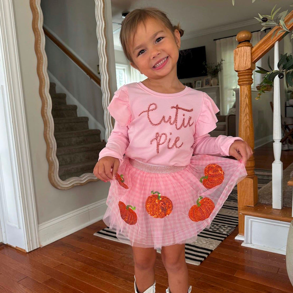 Smiling toddler in a pink shirt that says "Cutie Pie" and a pink tutu skirt with sequin pumpkins.