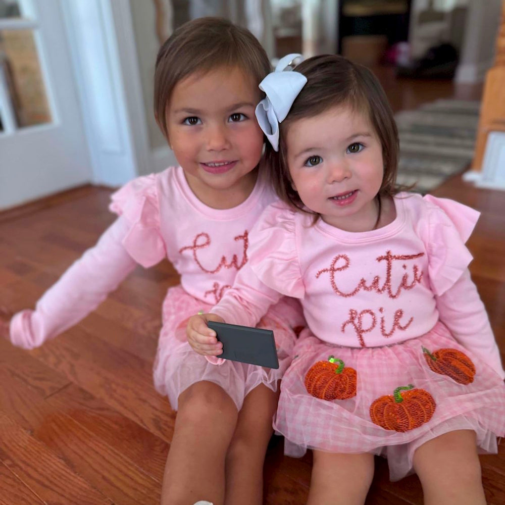 Two young girls smiling in matching pink "Cutie pie" shirts and sequined pumpkin skirts.