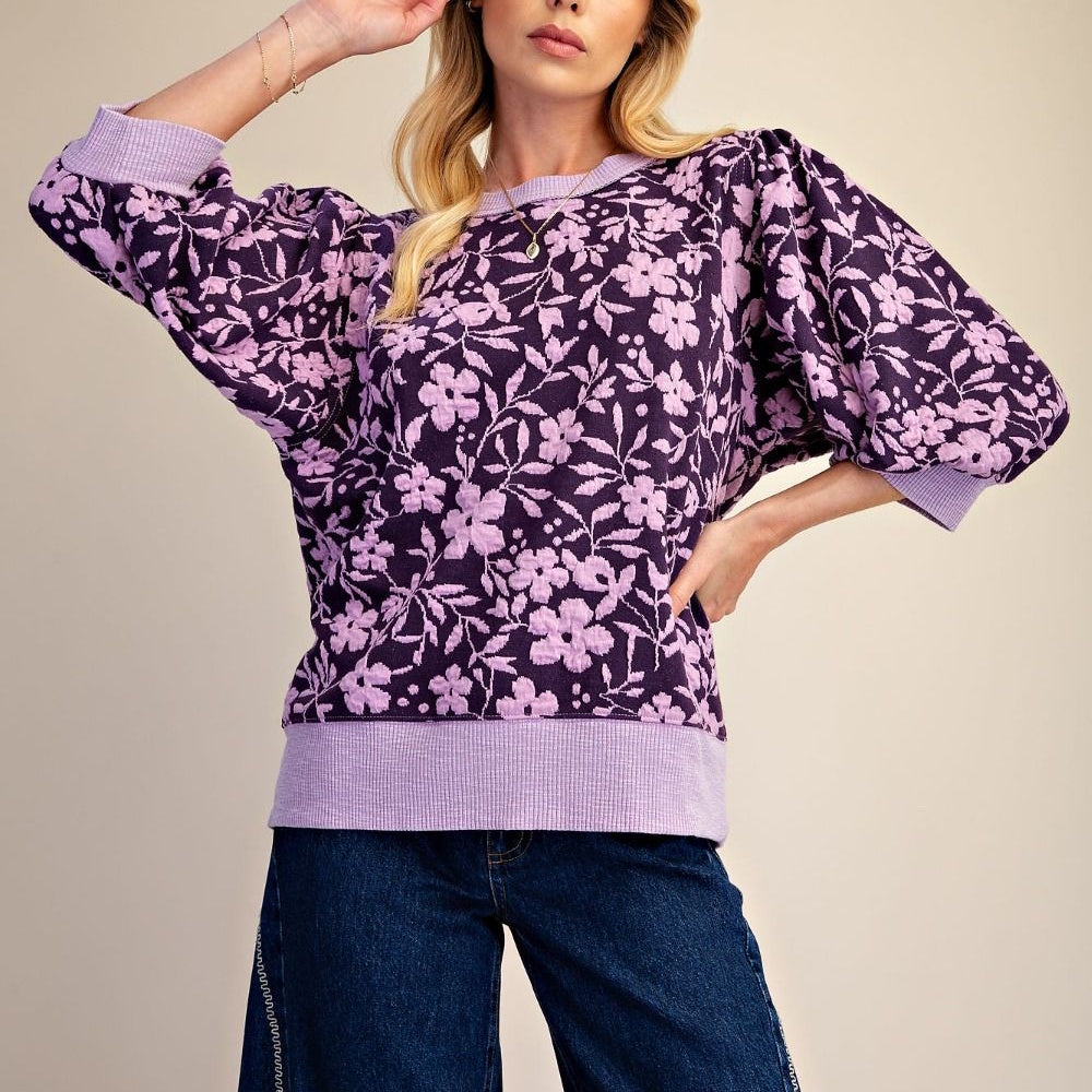 Woman wearing a purple floral-patterned puff-sleeve sweater with dark denim jeans.