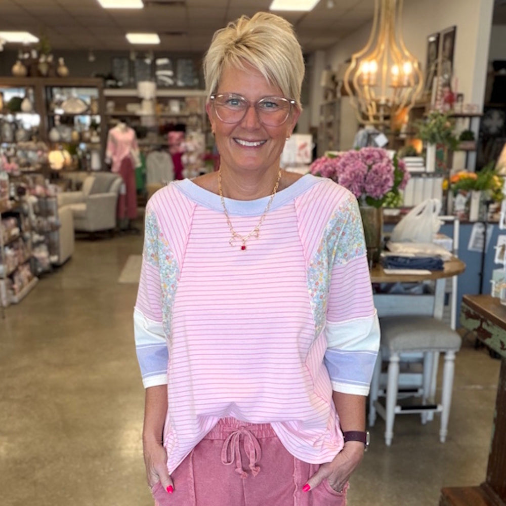 Woman in pink striped knit top with floral sleeve panels standing in a boutique shop.