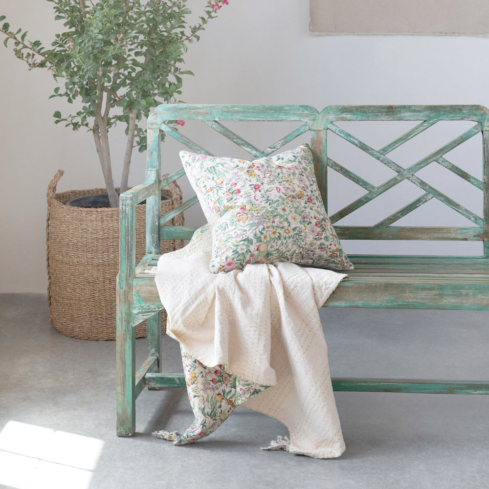 Floral cotton quilted throw and matching pillow on a rustic green wooden bench next to a potted plant in a woven basket.