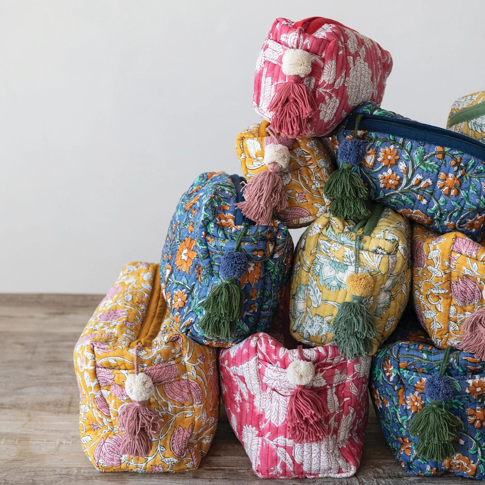 Stacked quilted cotton floral zip pouches in pink, blue, and yellow with decorative tassels on a wooden surface.