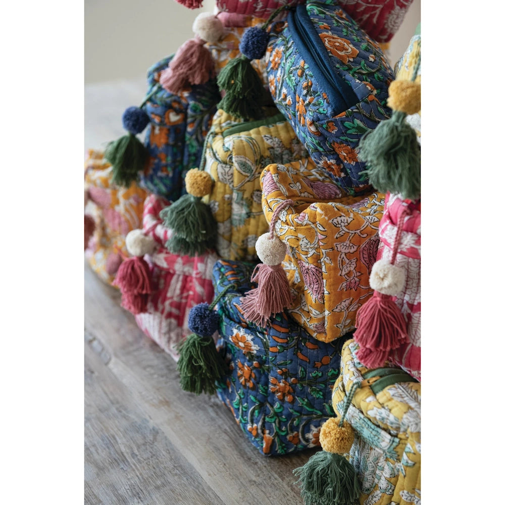 Stack of colorful quilted floral zip pouches in cotton voile with decorative yarn tassels on a wooden surface.