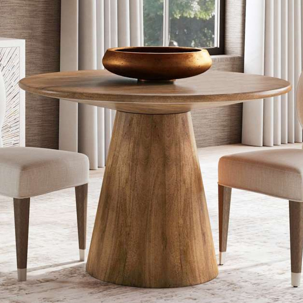 Modern round wood pedestal dining table with a bronze bowl centerpiece and upholstered chairs in a bright room.