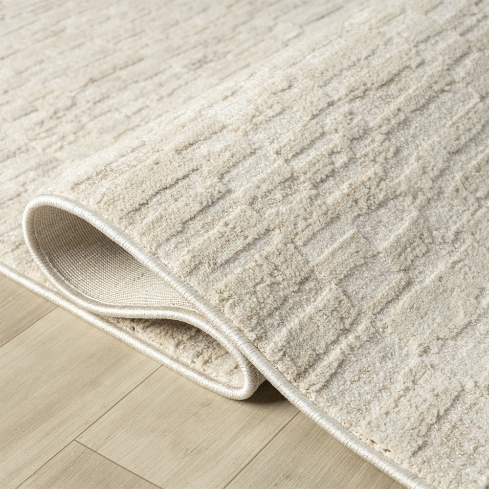 Close up of cream area rug corner showing soft textured pile, bound edges and subtle geometric pattern.