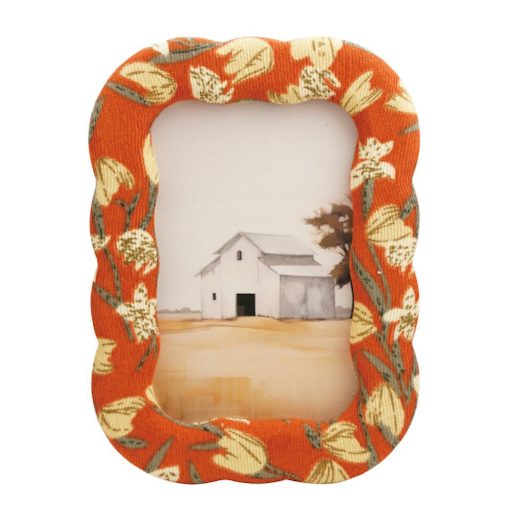 An orange fabric picture frame with a floral pattern, displaying a painting of a white rustic barn.
