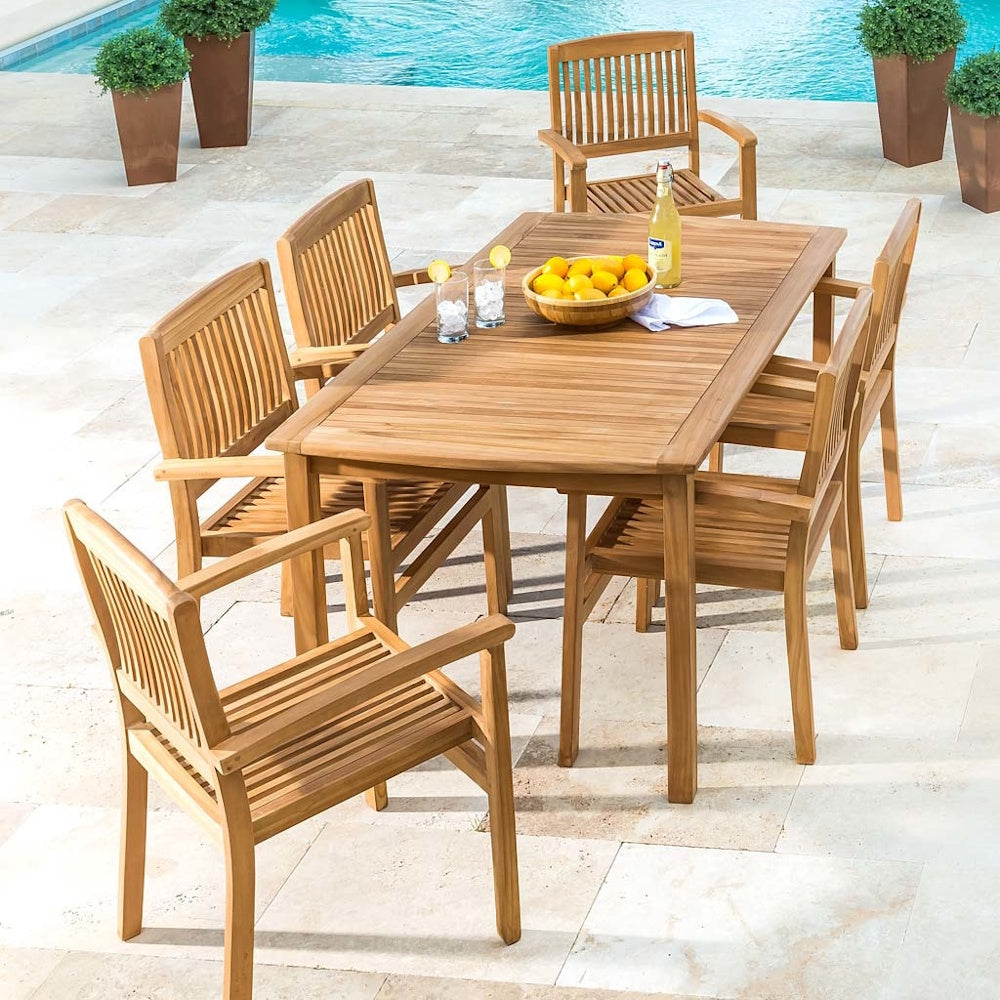 Outdoor teak dining table set with six slatted armchairs on a patio beside a swimming pool.