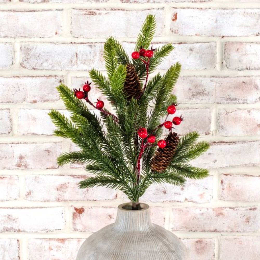 Faux evergreen spray with red berries and pinecones set in a vase against a white brick wall.