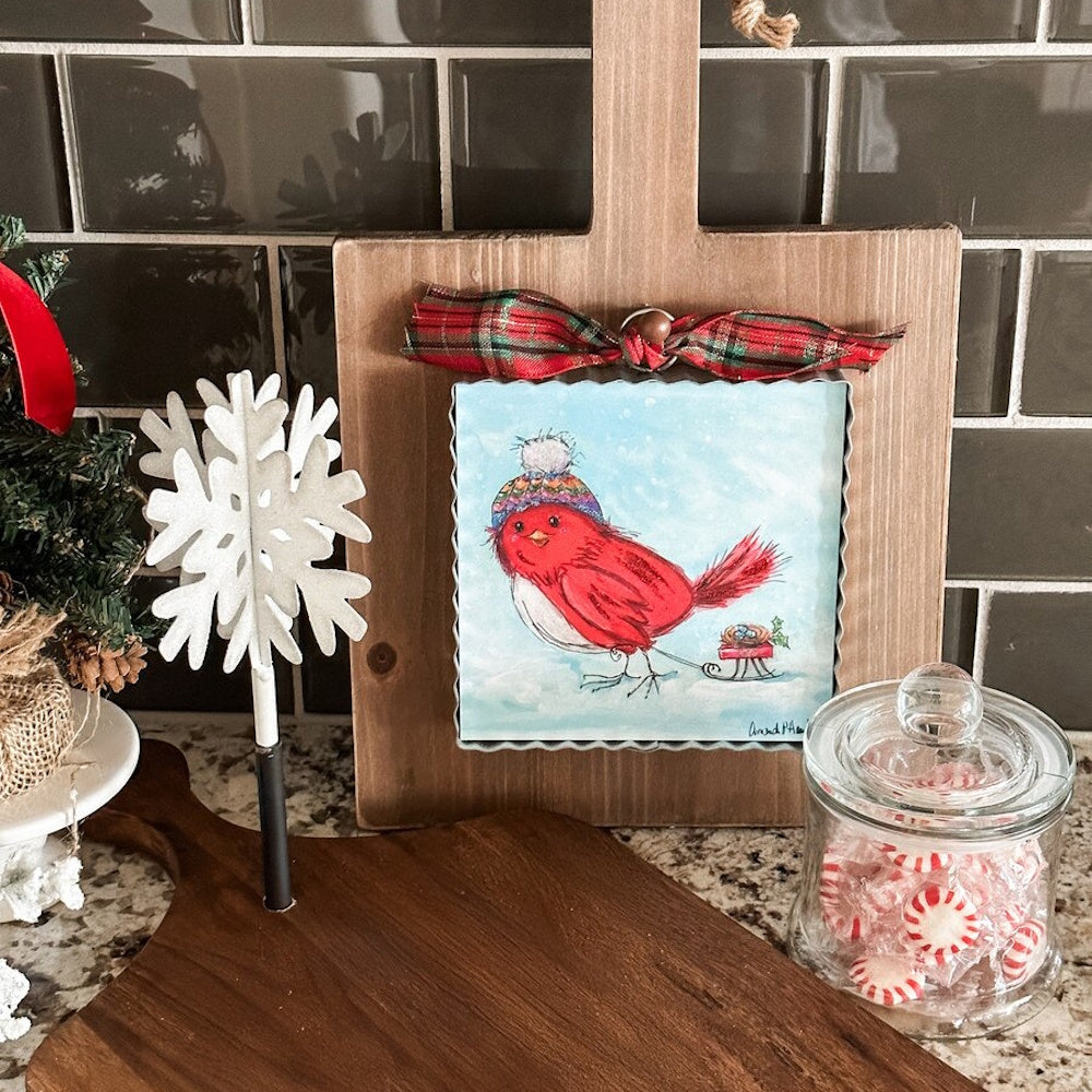 Front view of a square red bird metal art piece wearing a winter hat and pulling a sled.