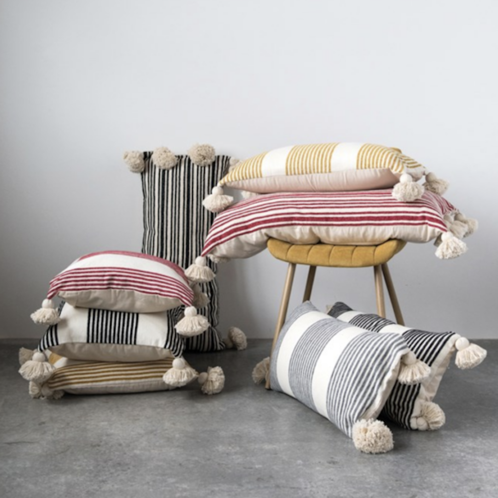 A stack and arrangement of striped throw pillows with tassels on a chair and floor against a plain wall.