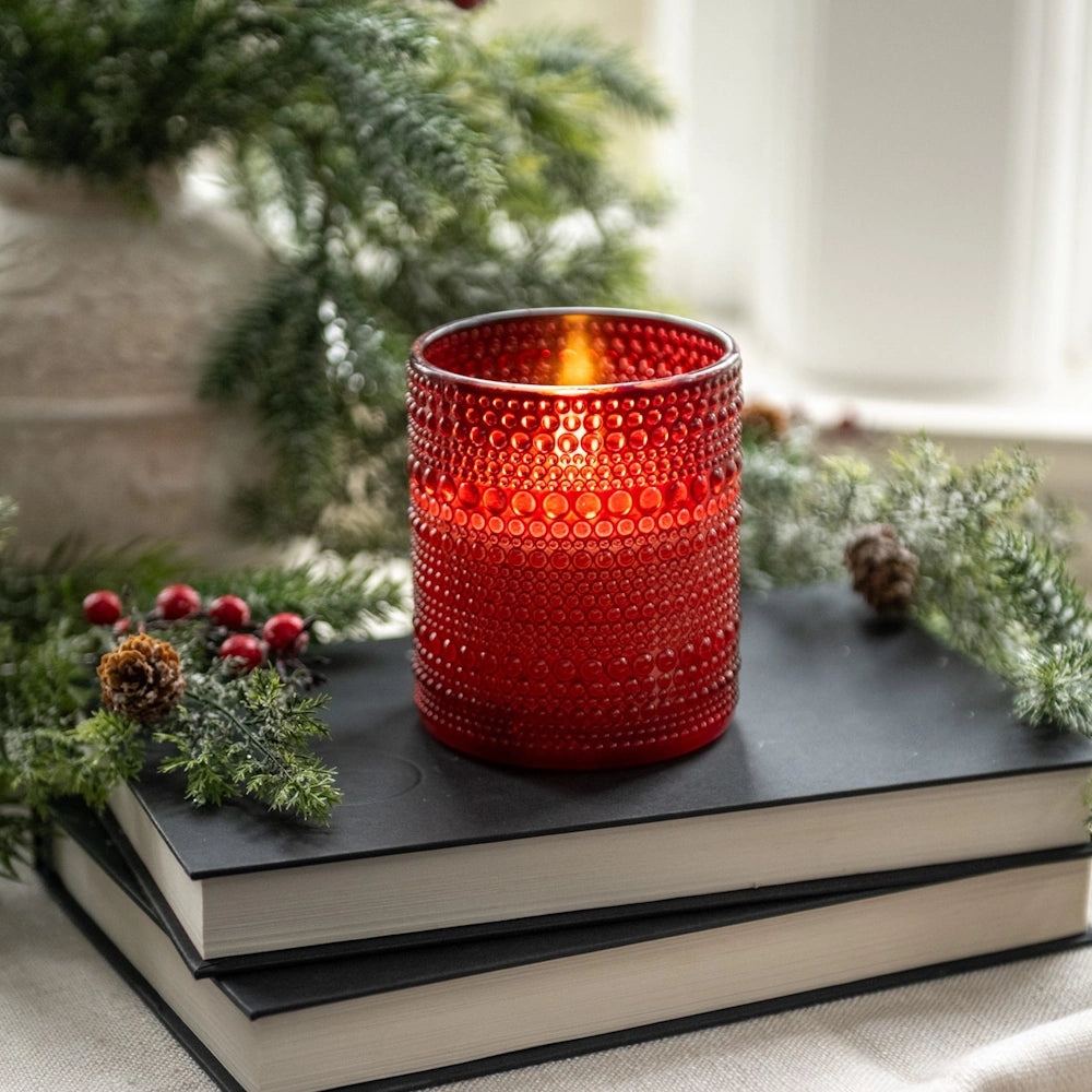 Red hobnail glass candle holder glowing on stack of books near festive pine greenery.