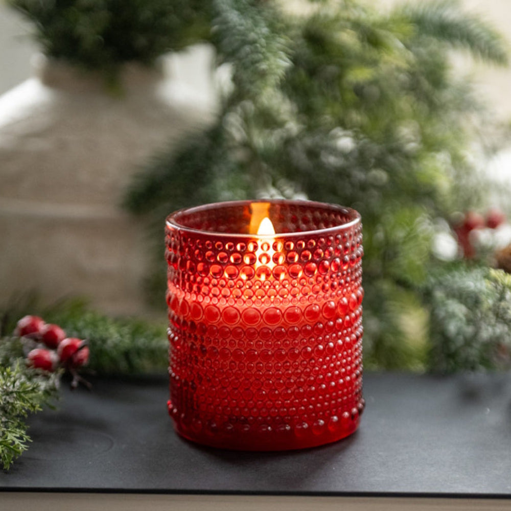 Red dotted glass votive with a glowing 3D flameless candle sitting on stacked books with pine greenery.