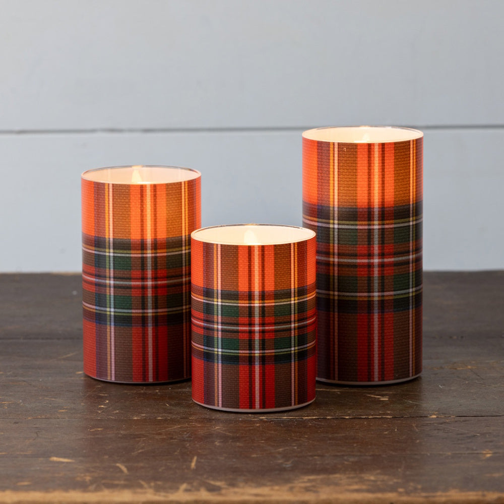 Three glowing red tartan plaid pillar candles of varying heights on a rustic wooden table.