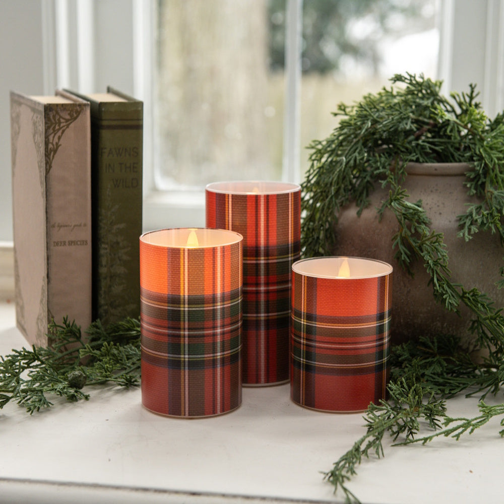 Three lit flameless pillar candles wrapped in red tartan plaid pattern by books and greenery.