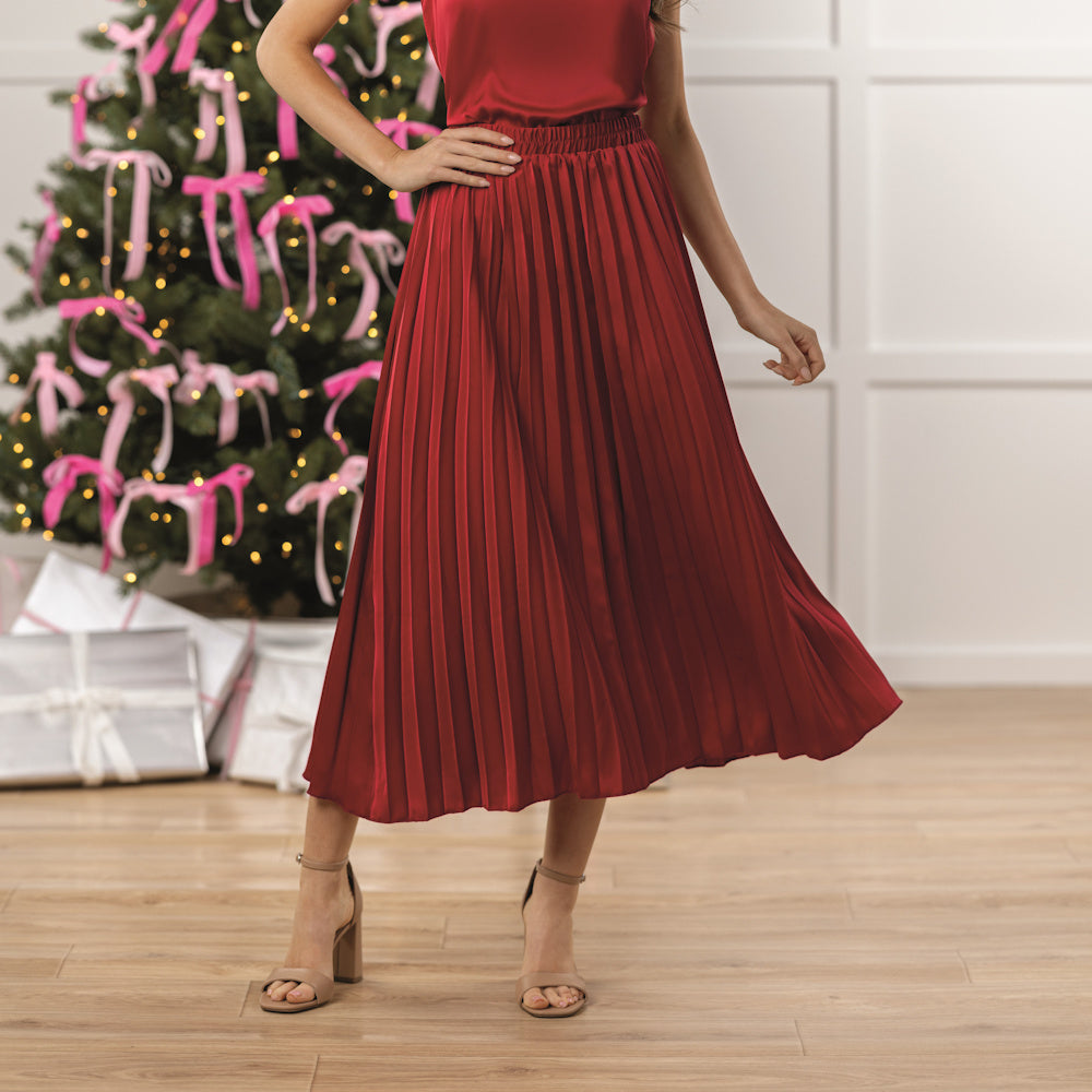 A woman in a red pleated skirt and top poses near a lit Christmas tree with pink bows and silver gifts.